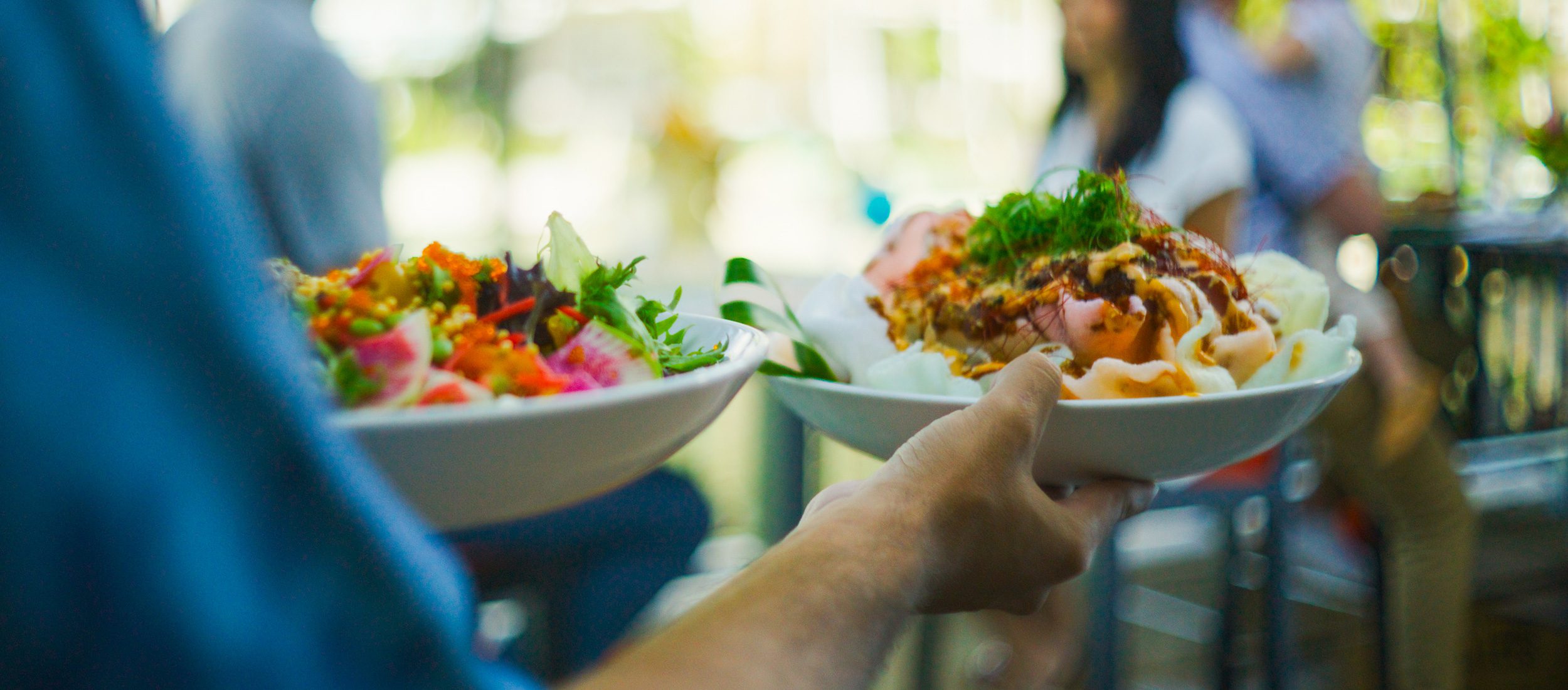 Couple dining at oceanfront restaurant with waiter holding two plates of food in the foreground.