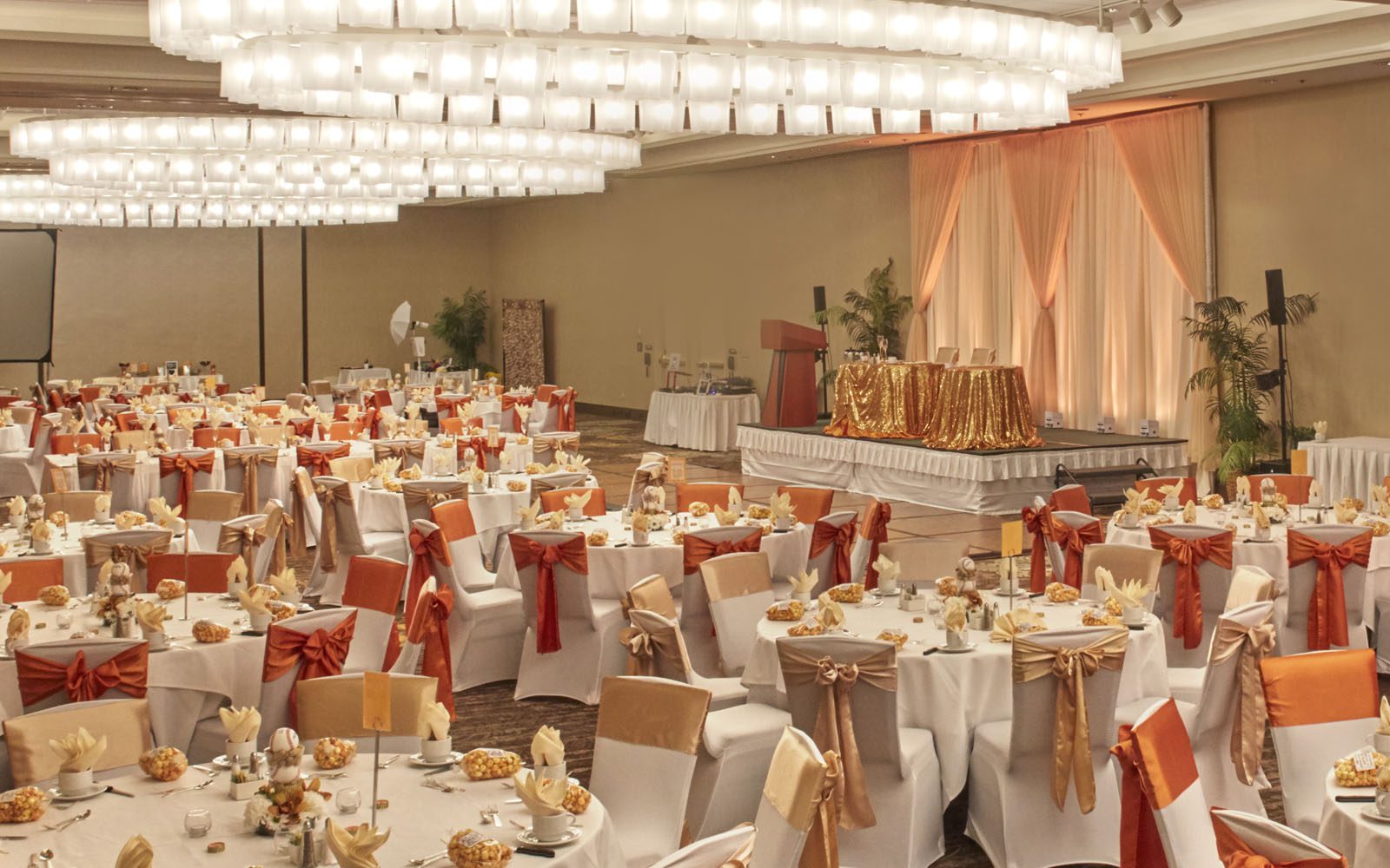 Weddings set up in the South Pacific Ballroom. Room is filled with white table clothed rounds and gold and orange chair ribbons with a gold stage in the background.