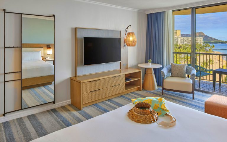 Rainbow Oceanfront King guest room with flat television, mirror and view of Diamond Head. A woven hat and beach bag sit on a white linen bed.