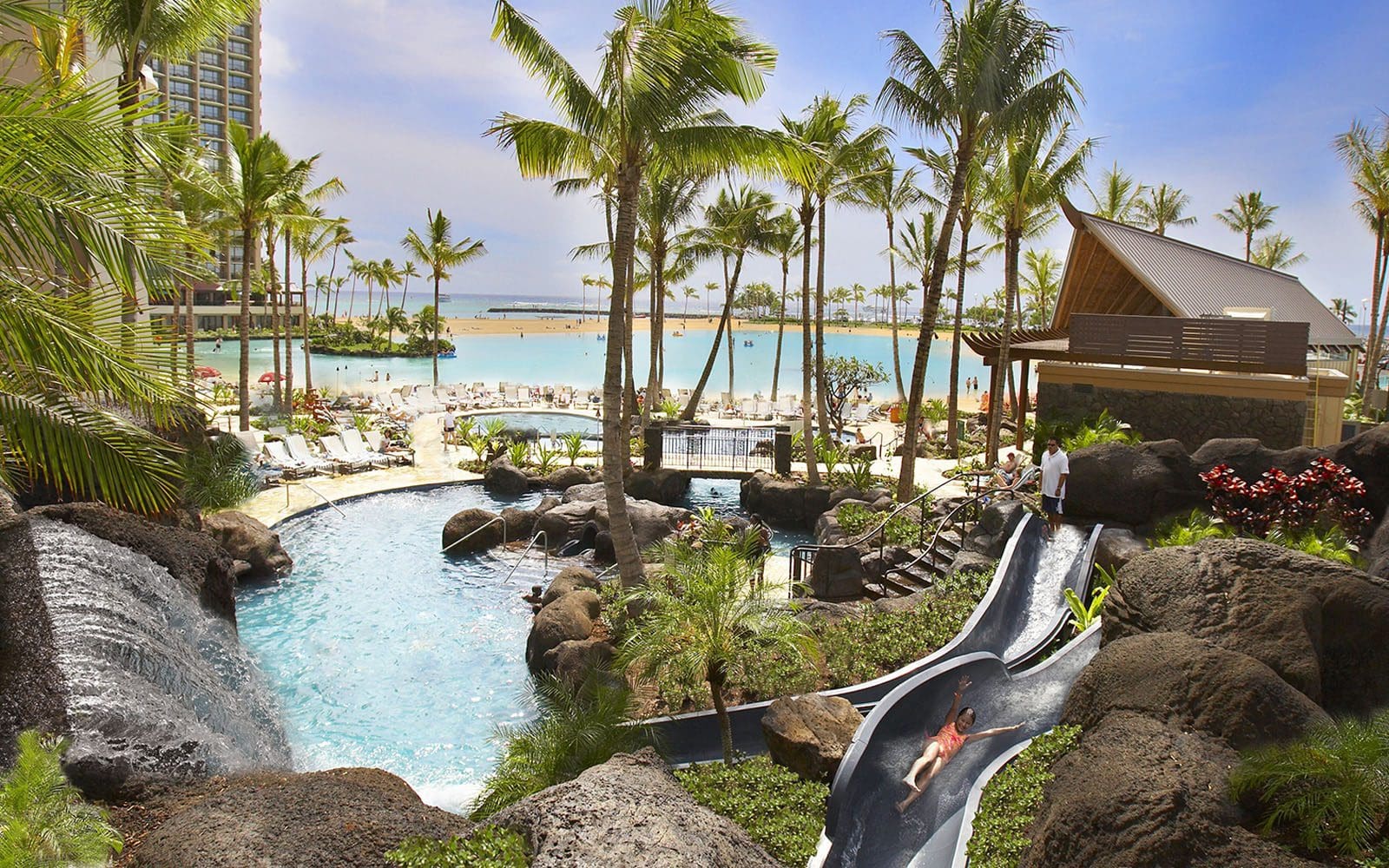 Paradise Pool with a girl going down the slide in the foreground and the lagoon and ocean in the background. Palm trees swaying.