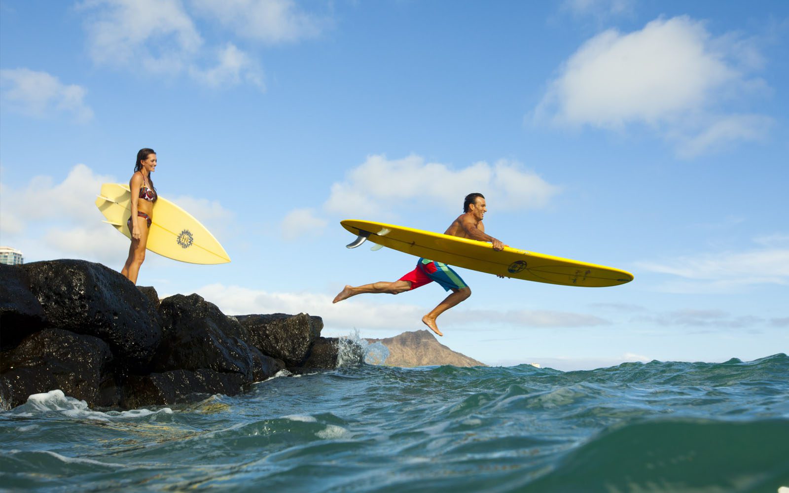 Couple jumping in to the ocean in front of Hilton Hawaiian Village with their surfboards